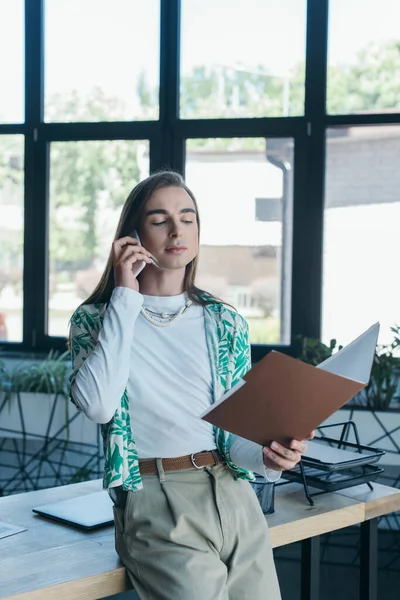 Queer designer holding paper folder and talking on smartphone in office - foto de stock