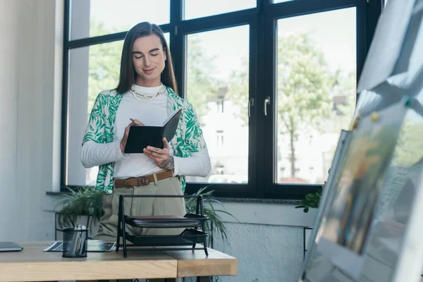 Positive queer designer writing on notebook in office — Stock Photo