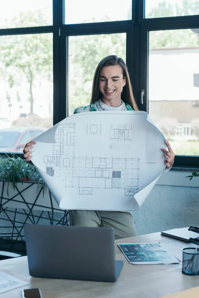 Smiling queer designer holding blueprint during video call on laptop in creative agency — Stock Photo