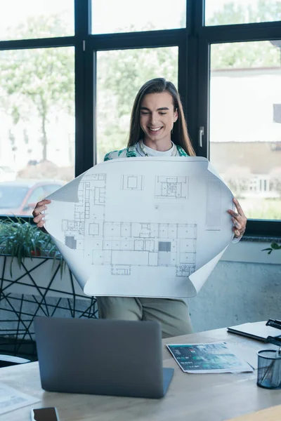 Smiling queer designer holding blueprint during video call on laptop in creative agency — Stock Photo