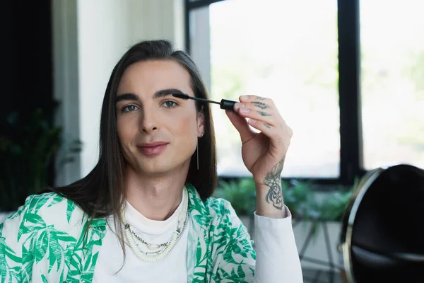 Queer designer applying mascara brush near blurred mirror in office — Foto stock