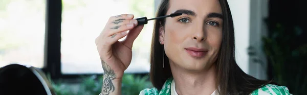 Nonbinary designer holding mascara brush and looking at camera in office, banner — Foto stock