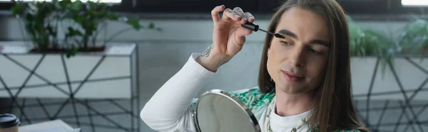 Nonbinary designer applying mascara near mirror in creative agency, banner - foto de stock