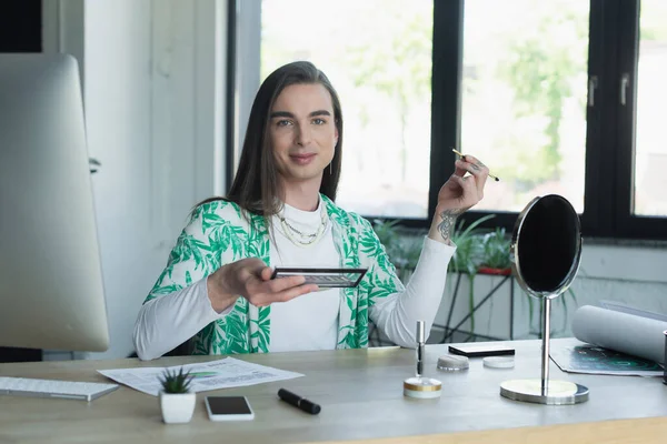 Smiling queer designer holding eye shadow near gadgets and mirror in office - foto de stock