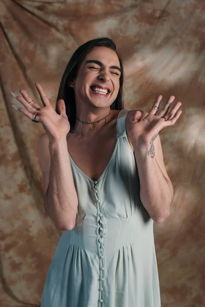 Весела татуйована небінарна особа в сукні закриває очі на абстрактному коричневому фоні — стокове фото