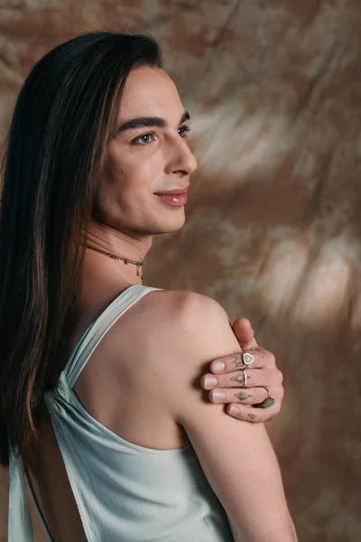 Young nonbinary person in dress touching shoulder on abstract brown background — Stockfoto