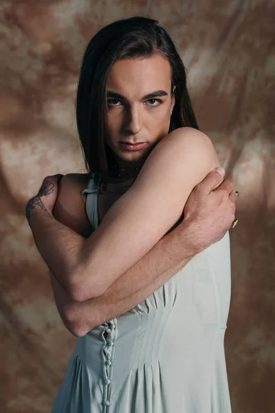 Portrait of nonbinary person in sundress touching shoulders on abstract brown background - foto de stock