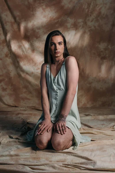 Young tattooed queer person looking at camera while sitting on abstract brown background - foto de stock