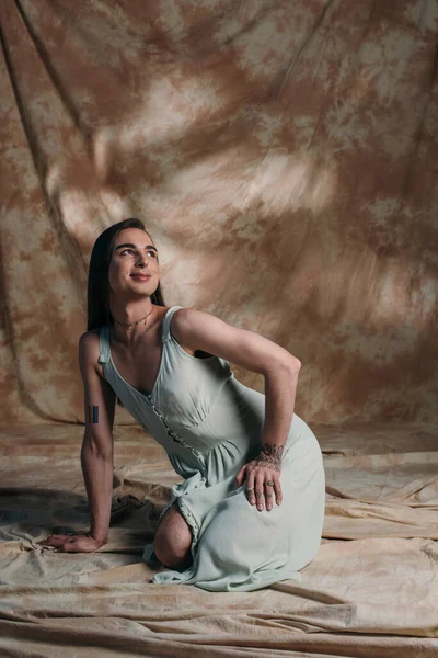 Smiling queer person in sundress looking away while sitting on abstract brown background - foto de stock