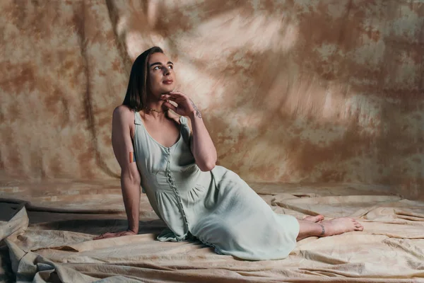 Queer person in sundress touching chin and looking away while sitting on abstract brown background — Stockfoto