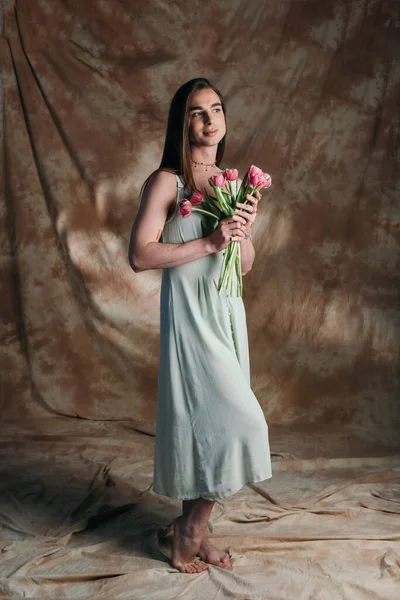 Full length of stylish queer person in sundress holding flowers on abstract brown background — Stockfoto