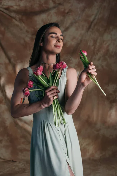 Young nonbinary person in sundress holding pink tulips on abstract brown background — Photo de stock