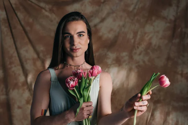 Brunette nonbinary person in dress holding bouquet and looking at camera on abstract brown background — Stock Photo