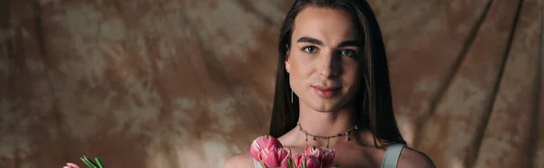 Young nonbinary person looking at camera near pink tulips on abstract brown background, banner — Foto stock