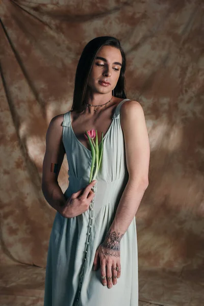 Young brunette queer person in dress holding pink tulip on abstract brown background — Fotografia de Stock