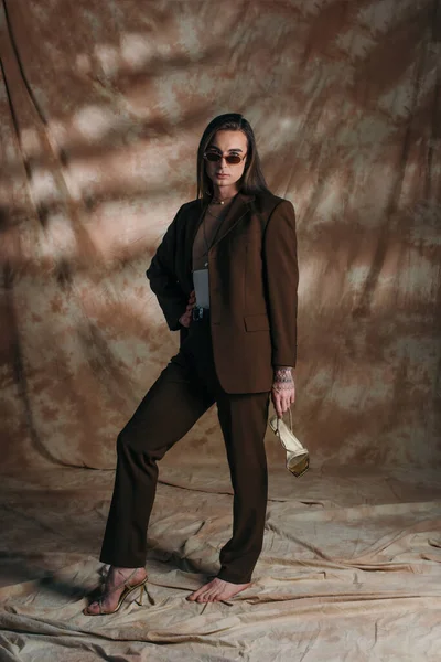 Модна небінарна особа в костюмі тримає взуття на абстрактному коричневому фоні — Stock Photo