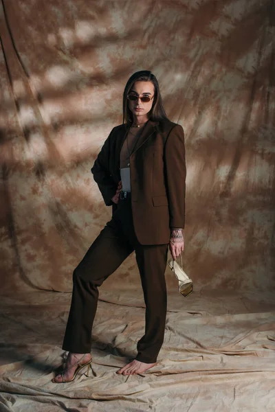 Trendy nonbinary person in suit holding shoe on abstract brown background — Stock Photo