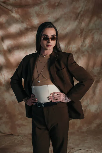 Brunette queer person in sunglasses holding hands on hips on abstract brown background — Stockfoto