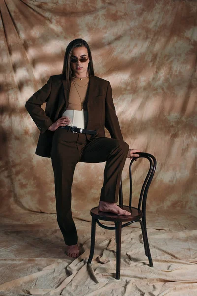 Стильна небінарна особа в костюмі та сонцезахисних окулярах позує біля стільця на абстрактному коричневому фоні — Stock Photo