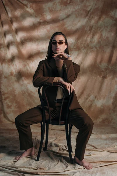 Barefoot queer person in sunglasses and suit sitting on chair on abstract brown background — Stockfoto