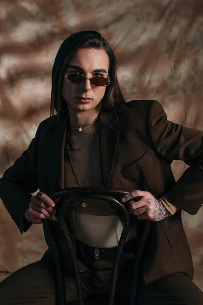 Портрет брюнетки в солнцезащитных очках и куртке, сидящей на стуле на абстрактном коричневом фоне — стоковое фото