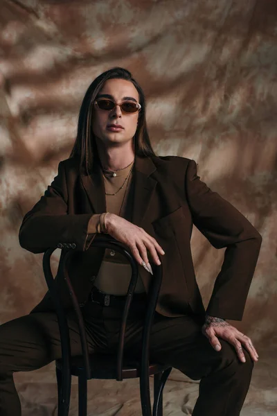 Stylish queer person in suit and sunglasses sitting on chair on abstract brown background - foto de stock