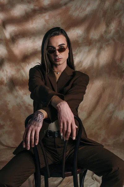 Fashionable tattooed queer person in sunglasses sitting on chair on abstract brown background — Foto stock