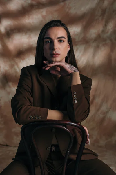 Queer person in jacket looking at camera while sitting on chair on abstract brown background — Stock Photo