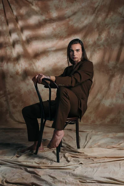 Barefoot queer person in suit posing on chair on abstract brown background — Stock Photo