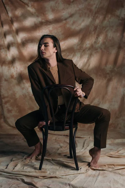 Barefoot queer person in blazer sitting on chair on abstract brown background — Stock Photo