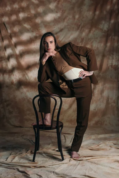 Brunette queer person in suit and corset posing near chair on abstract brown background - foto de stock