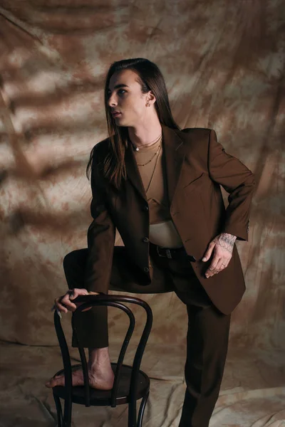 Trendy young nonbinary person in suit looking away near chair on abstract brown background — Stockfoto