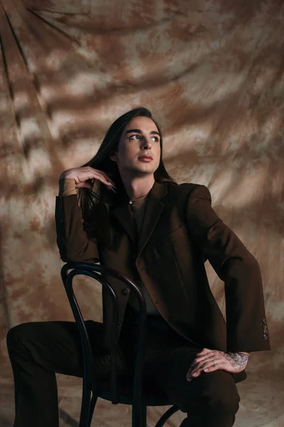 Trendy queer person in suit touching hair while sitting on chair on abstract brown background — Fotografia de Stock