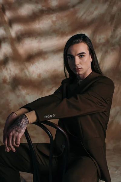 Portrait of trendy young nonbinary person sitting on chair on abstract brown background — Stockfoto