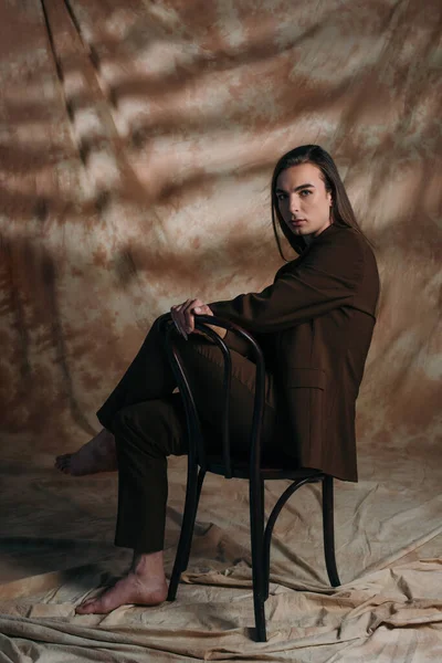 Полная длина брюнетки странный человек в костюме сидя на стуле на абстрактном коричневом фоне — стоковое фото