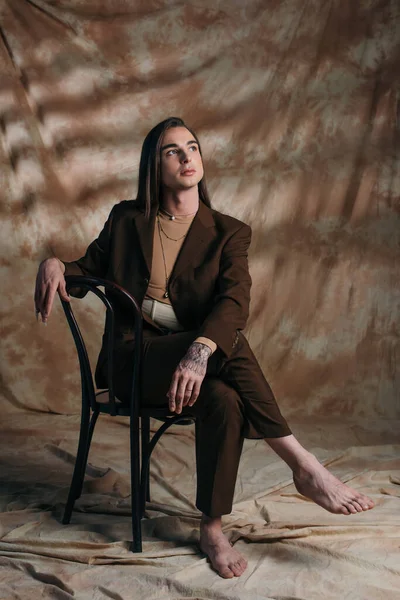 Barefoot nonbinary person in suit sitting on chair on abstract brown background — Fotografia de Stock