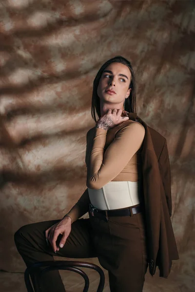 Nonbinary person in corset holding jacket near chair on abstract brown background — Stock Photo