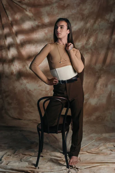 Trendy nonbinary person in corset holding jacket near chair on abstract brown background — Fotografia de Stock