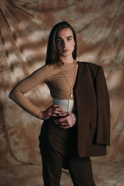 Brunette queer person in jacket holding hand on hip on abstract brown background — Fotografia de Stock