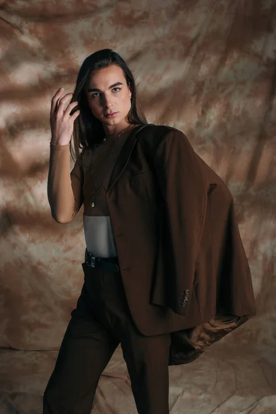 Queer person in blazer touching hair and looking at camera on abstract brown background — Foto stock