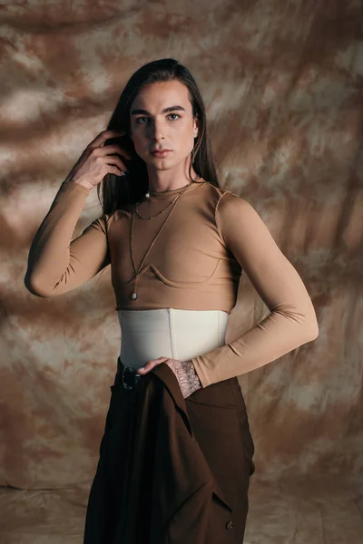 Stylish nonbinary person holding jacket and looking at camera on abstract brown background - foto de stock