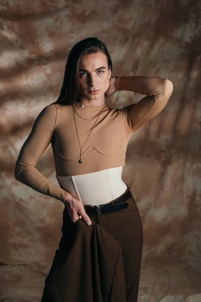 Trendy nonbinary person holding blazer and posing on abstract brown background — Stock Photo