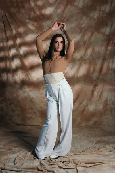 Stylish nonbinary person in corset and white pants standing on abstract brown background — Photo de stock