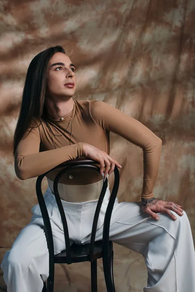 Brunette queer person in white pants sitting on chair on abstract brown background - foto de stock