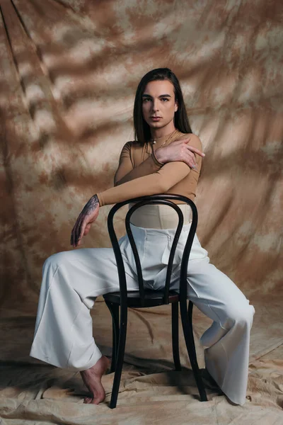 Barefoot nonbinary person looking at camera while sitting on chair on abstract brown background - foto de stock