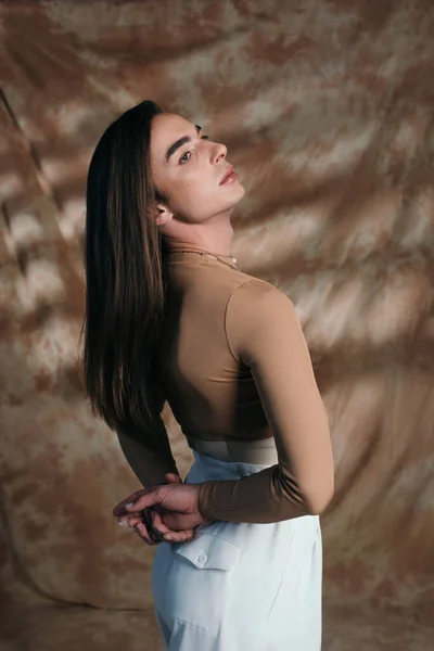 Young queer person looking away while standing on abstract brown background — Fotografia de Stock