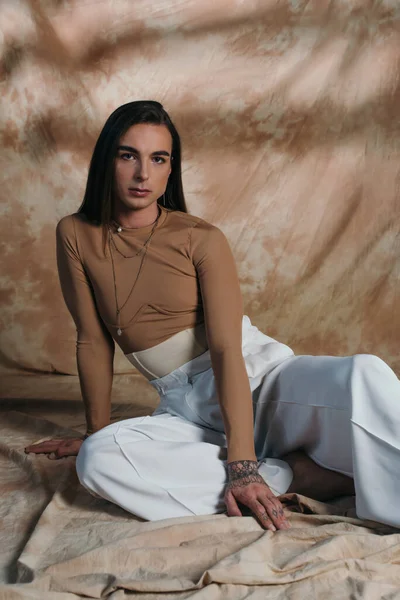 Stylish queer person in white pants sitting on abstract brown background — Photo de stock