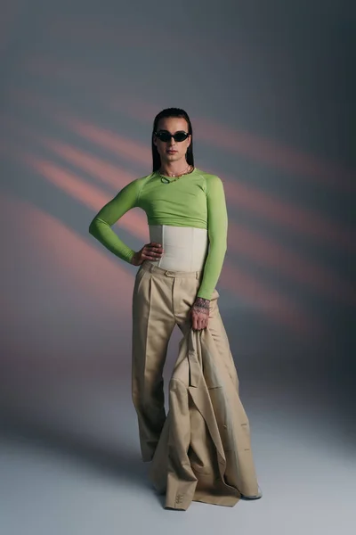 Full length of trendy nonbinary model in sunglasses holding jacket on abstract background — Foto stock