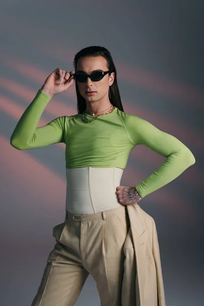Brunette nonbinary model in corset and sunglasses holding jacket on abstract background - foto de stock