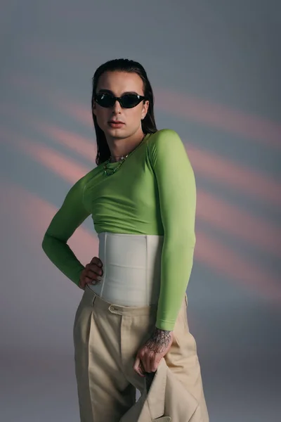 Fashionable nonbinary model in sunglasses holding jacket on abstract background — Photo de stock